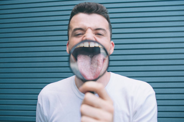 Funny man is standing and showing tongue through lenz. It is zoomed in. Guy is holding thing with one hand. Isolated on striped and blue background. - Foto, afbeelding
