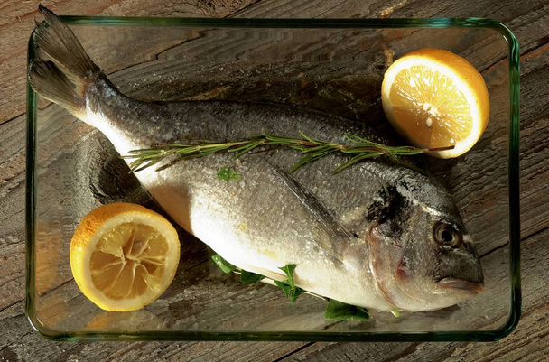 Έτοιμο για ψητό ψάρι Τσιπούρα με λεμόνια και το δενδρολίβανο στη μαγειρική δίσκος Γυάλινος κινηματογραφήσεων σε ξύλινο υπόβαθρο. Το Top View - Φωτογραφία, εικόνα