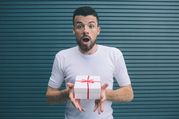 Very excited and surprised man is holding present in hands. He didnt expect to get it. Isolated on striped and blue background. - Foto, Imagen