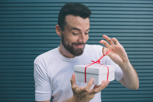 Delightful and nice guy is opening the present box. He is looking at irt and pulling out red ribbon. He is very excited. Isolated on striped and blue background. - Фото, изображение