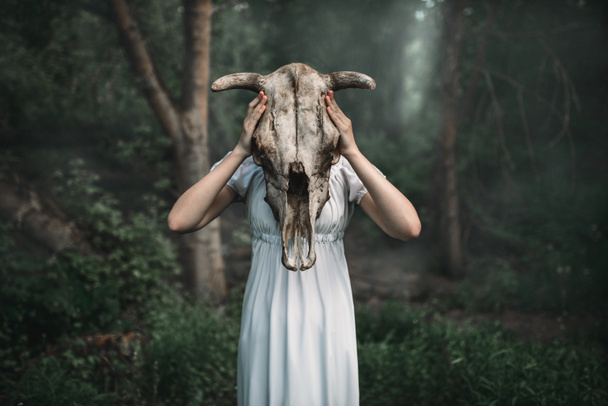 Γυναίκα θύμα σε λευκό φόρεμα με κρανίο του ζώου αντί για το κεφάλι, δάσους σε φόντο. Φωτογραφία σε στυλ τρόμου, εξορκισμός - Φωτογραφία, εικόνα