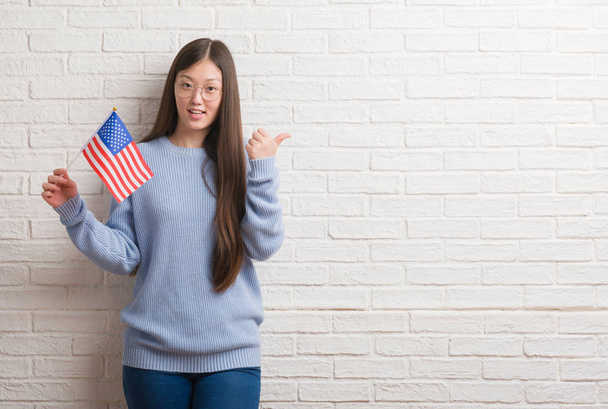 若い中国の女性アメリカの旗を保持しているレンガの壁を指していると幸せそうな顔を笑顔で側まで親指で示す - 写真・画像