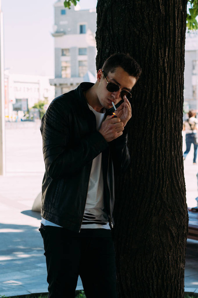 молодий хлопець курить біля дерева в місті. Чорна куртка, джинси, білі кросівки, біла футболка та окуляри. погана звичка. куріння в громадському місці
 - Фото, зображення