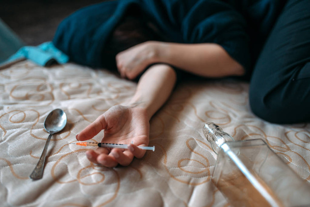 Vrouwelijke druggy met spuit in de hand ligt in bed, fles alcohol en lepel voor dosis voorbereiden op achtergrond. Drug verslaving concept, verslaafden - Foto, afbeelding