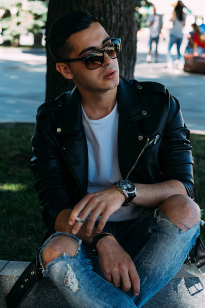 a young guy smokes near a tree in the city. A black jacket, jeans, white sneakers, a white T-shirt and glasses. bad habit. smoking in a public place - Photo, Image