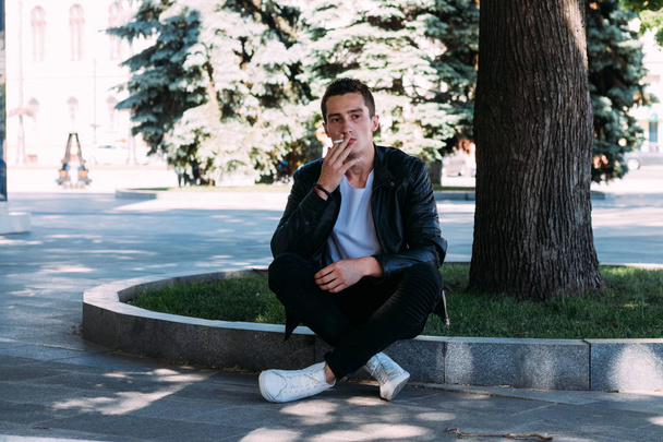 a young guy smokes near a tree in the city. A black jacket, jeans, white sneakers, a white T-shirt and glasses. bad habit. smoking in a public place - Fotografie, Obrázek