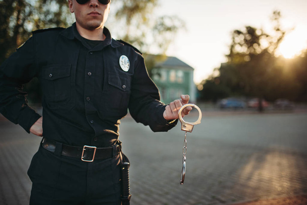Σοβαρή αξιωματικός της αστυνομίας στην στολή και γυαλιά ηλίου κατέχει χειροπέδες, θέα μπροστά. COP στο έργο. Έννοια δικαίου προστασίας, εργασία ελέγχου ασφαλείας - Φωτογραφία, εικόνα