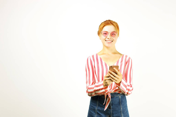 Young attractive natural redhead woman smiling at smartphone screen, using mobile phone, wearing knot shirt & cat eye glasses. White background, copy space. Female using online dating app concept. - Foto, Bild