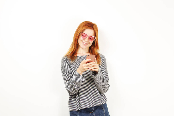 Young attractive natural redhead woman smiling at smartphone screen, using mobile phone, wearing loose grey shirt & cat eye glasses. White copy space background. Female using online dating app concept - Foto, immagini