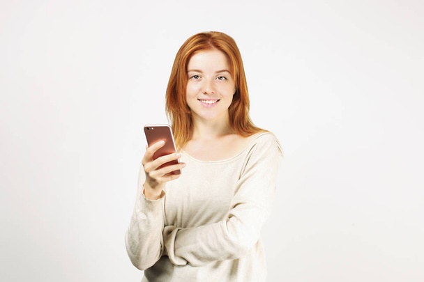 Young attractive natural redhead woman smiling at smartphone screen, using mobile phone, wearing loose white shirt. White background, copy space. Beautiful female using online dating app concept. - Фото, зображення