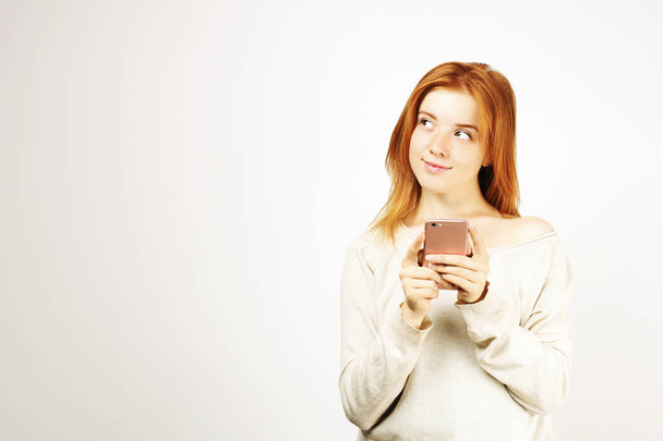 Young beautiful woman with long natural red hair, thinking facial expression and pose holding mobile cell phone. Attractive thoughtful hipster female with smartphone in hands. Background, copy space. - Photo, Image