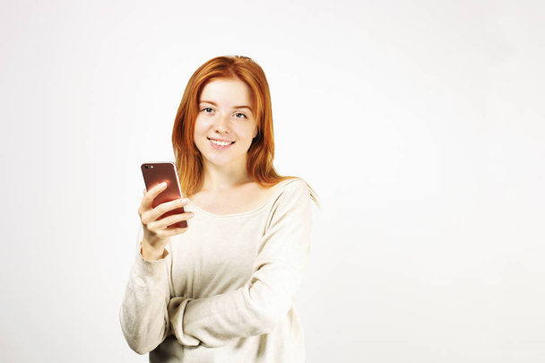 Young attractive natural redhead woman smiling at smartphone screen, using mobile phone, wearing loose white shirt. White background, copy space. Beautiful female using online dating app concept. - Photo, Image
