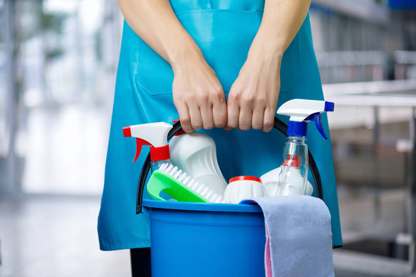 Limpiador con un cubo y productos de limpieza en un delantal se coloca sobre un fondo borroso
. - Foto, Imagen