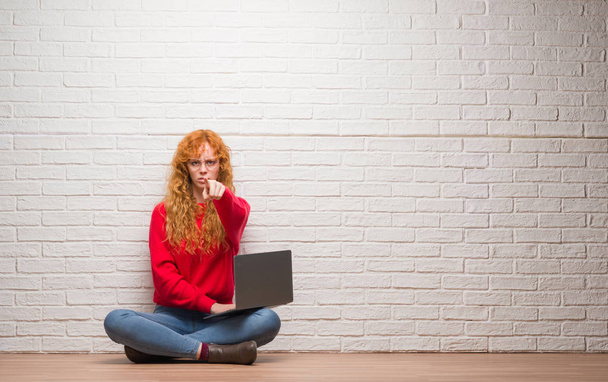 Νέοι κοκκινομάλλα γυναίκα κάθεται πάνω από τον τοίχο από τούβλα χρησιμοποιώντας υπολογιστή lap-top, δείχνοντας με το δάχτυλο στη φωτογραφική μηχανή και σε εσάς, σημάδι χέρι, θετική και σίγουρη κίνηση από το μέτωπο - Φωτογραφία, εικόνα