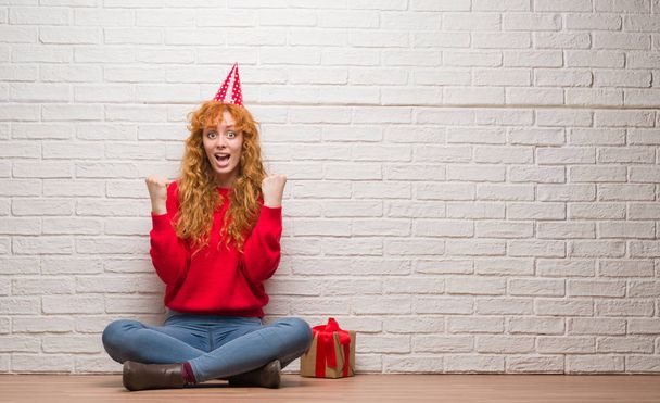 Nuori punapää nainen istuu tiiliseinällä yllään syntymäpäivä hattu huutaa ylpeä ja juhlii voittoa ja menestystä erittäin innoissaan, hurraavat tunteet
 - Valokuva, kuva