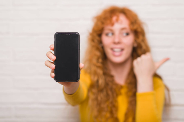 Jonge roodharige vrouw zitten over bakstenen muur tonen smartphone aan te wijzen en te tonen met de duim aan de kant met een blij gezicht lachend - Foto, afbeelding