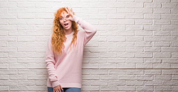 Young redhead woman standing over brick wall doing ok gesture shocked with surprised face, eye looking through fingers. Unbelieving expression. - Φωτογραφία, εικόνα