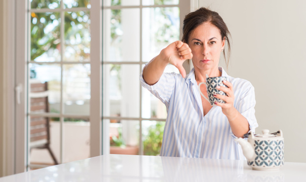 Mujer de mediana edad bebiendo una taza de té o café con la cara enojada, signo negativo que muestra aversión con los pulgares hacia abajo, concepto de rechazo
 - Foto, Imagen