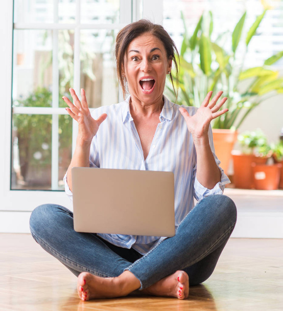 Frau mittleren Alters benutzt Laptop zu Hause sehr glücklich und aufgeregt, Siegerausdruck feiert Sieg schreiend mit breitem Lächeln und erhobenen Händen - Foto, Bild