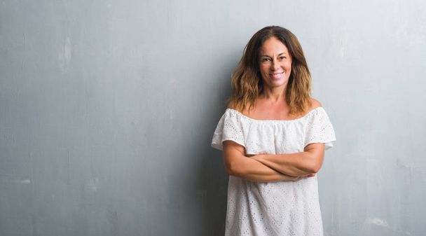 Eine hispanische Frau mittleren Alters steht über einer grauen Grunge-Wand und lächelt mit verschränkten Armen in die Kamera. positive Person. - Foto, Bild