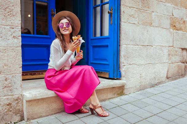 Young stylish woman eating fastfood on city street. Happy girl having tasty stuffed pancake outdoors. Summer holidays - Foto, Imagem