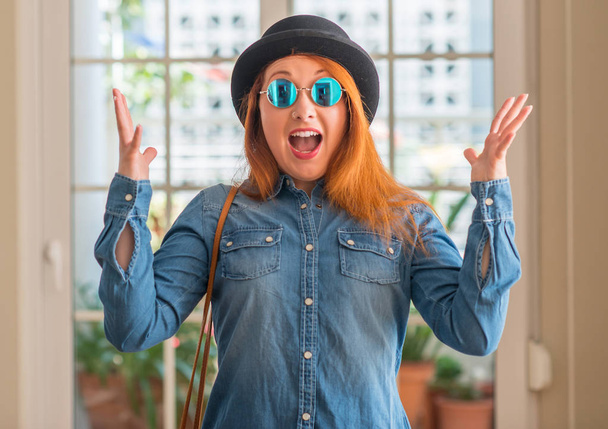 Femme rousse élégante portant un chapeau melon et des lunettes de soleil célébrant fou et étonné pour le succès avec les bras levés et les yeux ouverts hurlant excités. Concept gagnant
 - Photo, image