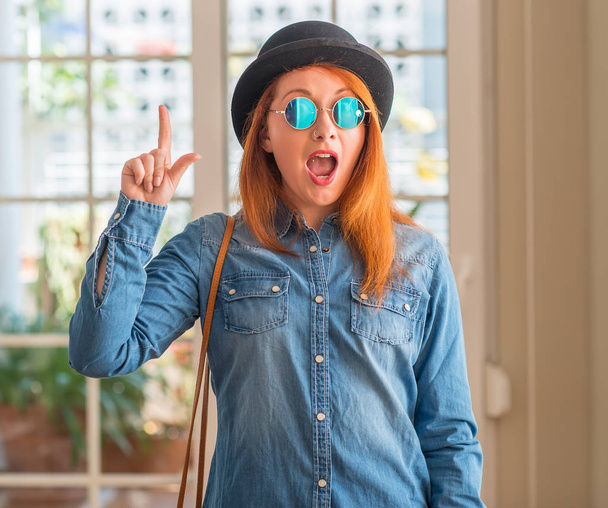 Стильная рыжая женщина в кепке и солнцезащитных очках, указывающих пальцем на успешную идею. Взволнован и счастлив. Номер один
. - Фото, изображение