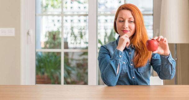 Redhead woman holding red apple at home serious face thinking about question, very confused idea - Foto, imagen