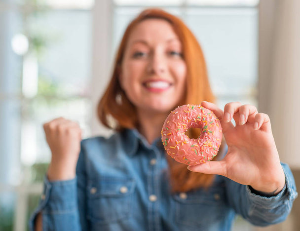 Redhead woman holding donut at home screaming proud and celebrating victory and success very excited, cheering emotion - Photo, Image