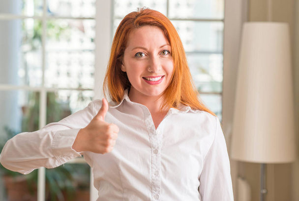 Рыжая женщина в белой рубашке дома счастлива с большой улыбкой делает хорошо знак, большой палец вверх пальцами, отличный знак
 - Фото, изображение
