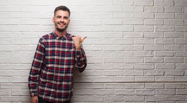 Νεαρός ενήλικος πάνω από το λευκό τοίχο τοποθετώντας και δείχνει με αντίχειρα μέχρι την πλευρά με το χαρούμενο πρόσωπο χαμογελά - Φωτογραφία, εικόνα