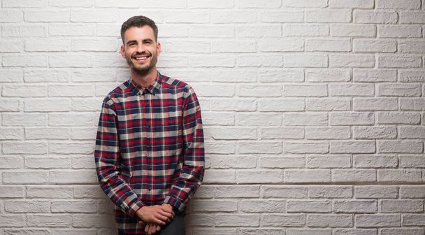 Молодий дорослий чоловік над білою цегляною стіною з щасливим обличчям, що стоїть і посміхається з впевненою посмішкою, показуючи зуби
 - Фото, зображення