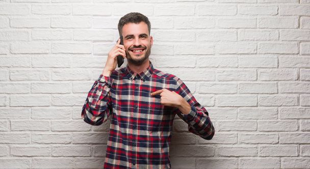 Молодой взрослый мужчина говорит по телефону, стоя над белой кирпичной стеной с удивленным лицом, указывающим пальцем на себя
 - Фото, изображение