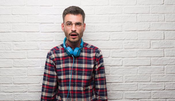 Young adult man over brick wall wearing headphones scared in shock with a surprise face, afraid and excited with fear expression - Photo, Image
