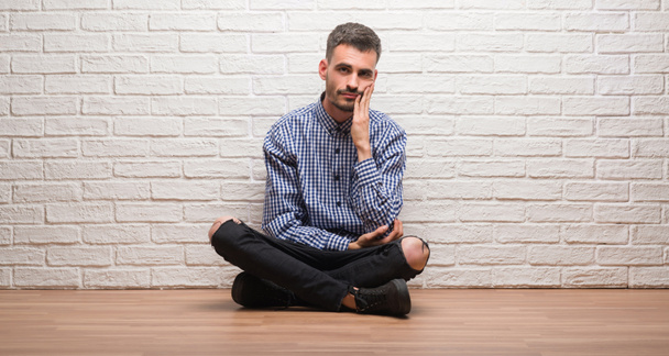 Νεαρός ενήλικος κάθεται πάνω από το λευκό τούβλο τοίχο σκέψης αναζητούν κουρασμένος και βαρεθεί με προβλήματα κατάθλιψης με σταυρωμένα χέρια. - Φωτογραφία, εικόνα