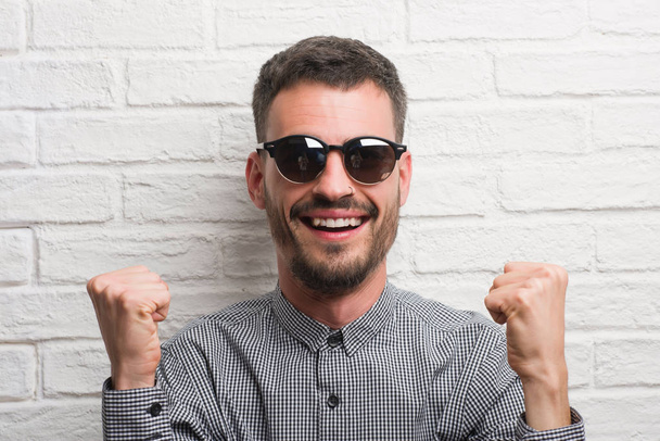 Hombre adulto joven con gafas de sol de pie sobre la pared de ladrillo blanco gritando orgulloso y celebrando la victoria y el éxito muy emocionado, animando la emoción
 - Foto, Imagen