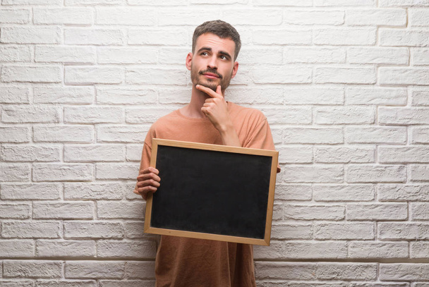 Jonge volwassen man over bakstenen muur holding schoolbord ernstige gezicht na te denken over de vraag, zeer verward idee - Foto, afbeelding