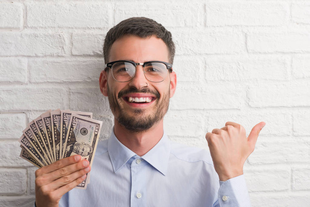Молодой бизнесмен-хипстер, держащий в руках доллары, показывая пальцем вверх в сторону, улыбаясь счастливым лицом
 - Фото, изображение