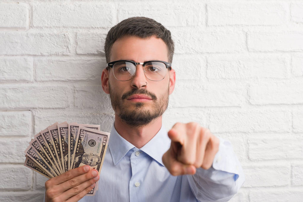 Young hipster business man holding dollars pointing with finger to the camera and to you, hand sign, positive and confident gesture from the front - 写真・画像