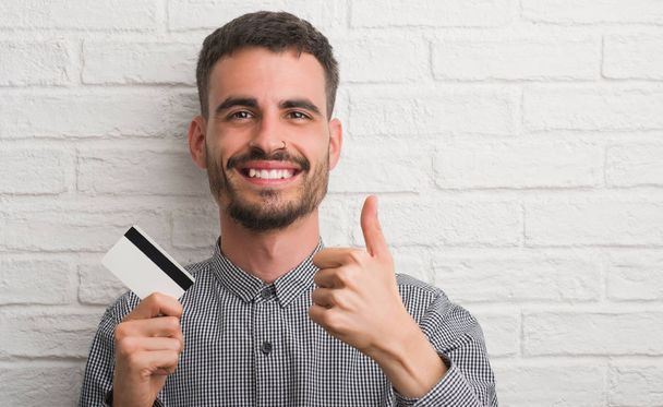 Jeune homme adulte sur le mur de briques tenant la carte de crédit heureux avec un grand sourire faisant signe ok, pouce levé avec les doigts, excellent signe
 - Photo, image