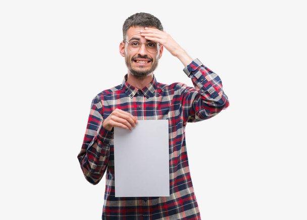 Joven hombre adulto hipster sosteniendo hoja de papel en blanco estresado con la mano en la cabeza, conmocionado con vergüenza y cara sorpresa, enojado y frustrado. Miedo y malestar por error
. - Foto, Imagen