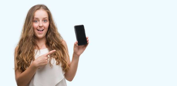 Νεαρή ξανθιά γυναίκα χρησιμοποιώντας smartphone πολύ χαρούμενος, δείχνοντας με το χέρι και το δάχτυλό - Φωτογραφία, εικόνα