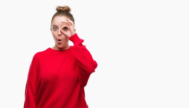 Młoda kobieta blonde noszenie bun i czerwony sweter robi ok gest wstrząśnięty z zaskoczony twarz, oczy, patrząc przez palce. Niewierzący wyrażenie. - Zdjęcie, obraz