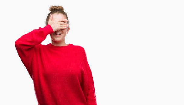 Young blonde woman wearing bun and red sweater smiling and laughing with hand on face covering eyes for surprise. Blind concept. - Foto, Bild