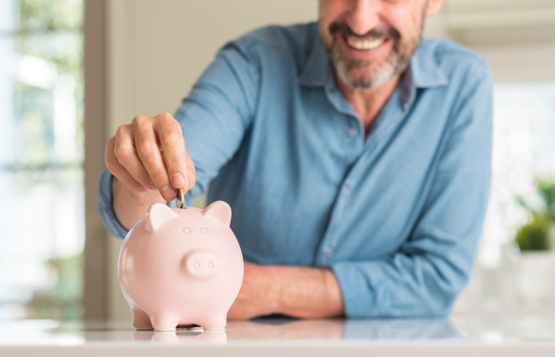 Orta yaş adam ayakta ve diş gösterilen kendine güvenen bir gülümsemeyle gülümseyerek mutlu bir yüz ile kumbara üzerinde para tasarrufu - Fotoğraf, Görsel