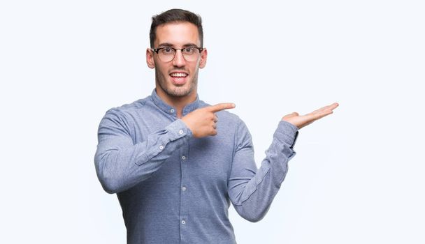 Handsome young elegant man wearing glasses amazed and smiling to the camera while presenting with hand and pointing with finger. - Photo, Image