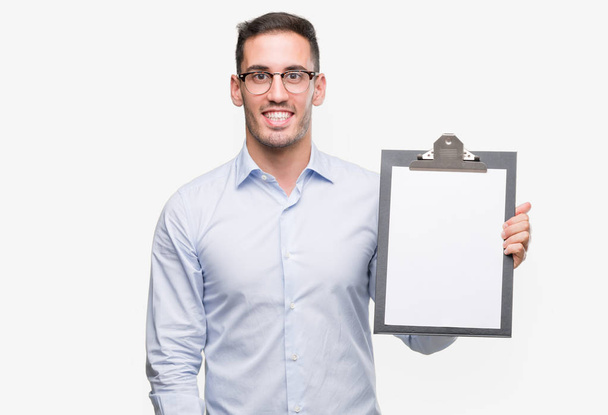 Handsome young business man showing a clipboard with a happy face standing and smiling with a confident smile showing teeth - Foto, Imagen