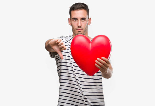 Pohledný mladý muž v lásce držící červené srdce s naštvaný obličej, záporné znaménko ukazující nechuť s palci dolů, koncept odmítnutí - Fotografie, Obrázek