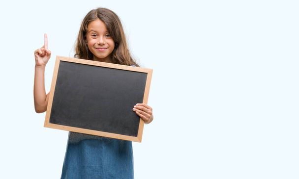 Brunette hispanic girl holding blackboard surprised with an idea or question pointing finger with happy face, number one - Foto, afbeelding
