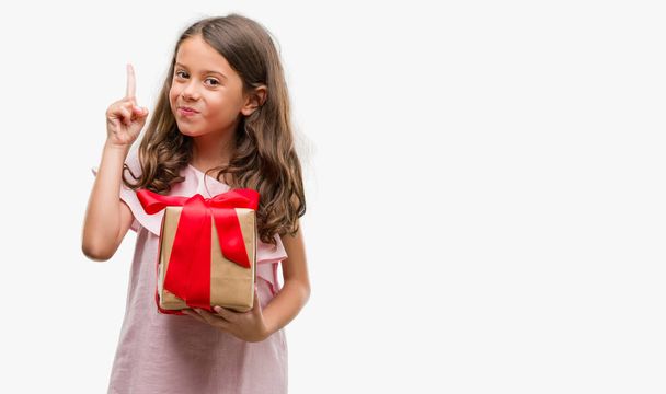 Brunette hispanic girl holding a gift surprised with an idea or question pointing finger with happy face, number one - Photo, Image